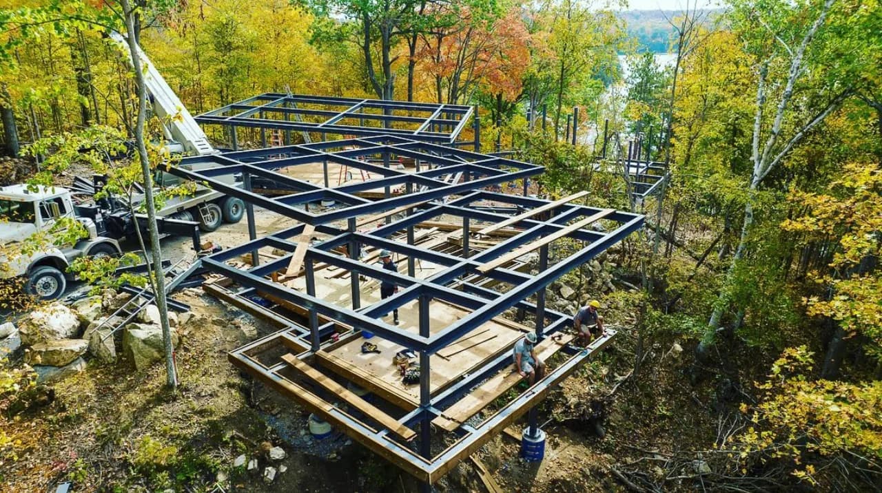 Installed black structural steel frame on a wooded construction site with field crew and crane access